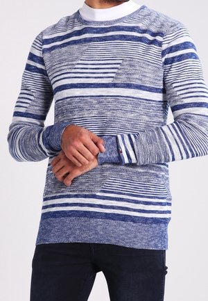 Mężczyzna w niebiesko-białym, paskowanym, dzianinowym swetrze z geometrycznymi wzorami, stoi ze splecionymi rękami z przodu, ubrany w ciemne spodnie.