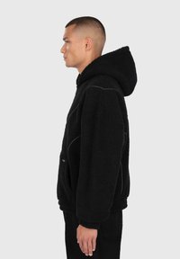 Giovane uomo con capelli corti che indossa un hoodie in pile nero con un ampio cappuccio, rivolto verso sinistra su uno sfondo chiaro e semplice.