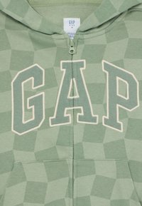 Felpa verde con zip caratterizzata da un motivo geometrico, logo "GAP" in crema e tasca frontale. Realizzata in tessuto morbido e con cappuccio.