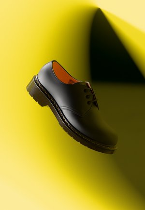 Schwarzer Lederschuh mit runder Spitze und vier Ösen. Verfügt über eine strukturierte Sohle und ein oranges Innenfutter. Vor einem gelben Hintergrund platziert.