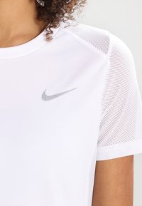 Camisa atlética blanca con mangas de malla y un logotipo de Nike en gris en el pecho, llevada por una persona con cabello rizado.