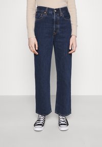 Jeans de mezclilla azul oscuro con un corte holgado, cintura alta y piernas rectas. Cuenta con cinco bolsillos y costuras en contraste. Se llevan con zapatillas negras.