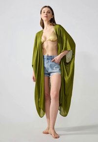 Copa de kimono fluido en verde oliva, con mangas sueltas, combinado con un bikini dorado y unos pantalones cortos de mezclilla. Tela suave con un aspecto texturizado.