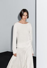 Lichtgewicht beige trui met een boothals en lange mouwen, gecombineerd met een vloeiende beige rok, tegen een neutrale achtergrond.