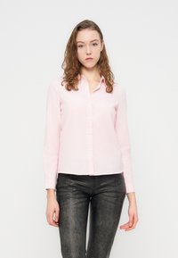 Πουκάμισο με μακριά μανίκια και κουμπιά σε ανοιχτό ροζ βαμβάκι με γιακά, διαθέτει ίσια ραφή στο κάτω μέρος και άνετη εφαρμογή. Φορεμένο με παντελόνια σκούρου χρώματος με υφή.