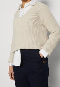 Beige gebreide trui met een V-hals, gedragen over een wit overhemd met kraag. De trui heeft een geribbelde structuur en een losse pasvorm.