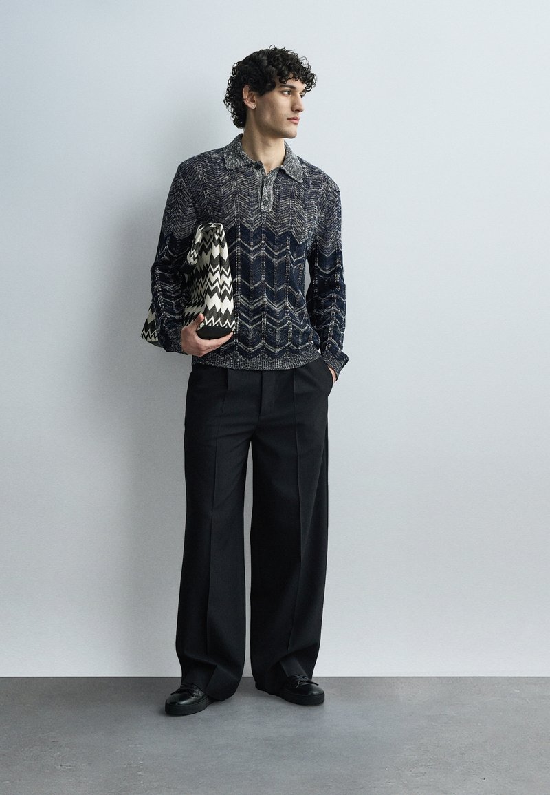 Man stands against gray wall wearing patterned dark sweater, wide black trousers, black shoes, holding large zigzag-patterned black and white bag.