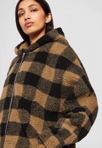 Beige und schwarze karierte Fleecejacke mit Reißverschluss, großem Kapuze und strukturierter Oberfläche. Verfügt über eine lässige Passform und weiches, gemütliches Material.