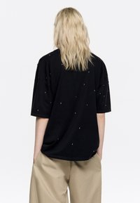 Camiseta negra de gran tamaño con tachuelas plateadas dispersas, mangas cortas y un cuello redondo. La tela parece suave y ligeramente texturizada.