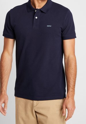 Mand iført en marineblå Esprit polo-shirt med beige bukser, stående med armene afslappet langs siden mod en ensfarvet baggrund.