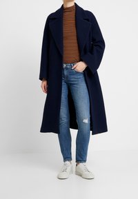 Abrigo largo azul marino con un cuello estructurado, llevado sobre una blusa de rayas marrón y negra. Combinado con jeans desgastados de color azul claro y zapatillas blancas.