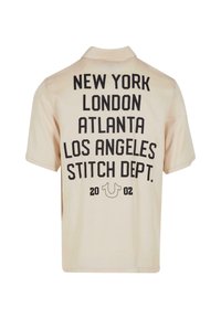 Polo de manga corta beige con texto negro: "NUEVA YORK LONDRES ATLANTA LOS ÁNGELES DEPARTAMENTO DE COSIDO 2002" en la parte posterior. Material de algodón suave.