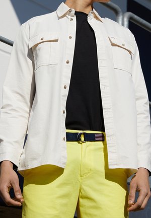 Osoba ubrana w białą koszulę na guziki na czarną koszulkę, jasnopomarańczowe spodnie i granatowy pasek z paskowaną klamrą.