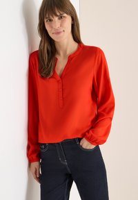 Blusa rossa a maniche lunghe con scollo a V bottonato, dotata di polsini arricciati e una superficie liscia, abbinata a jeans scuri.