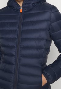 Marineblå pufferjakke med horisontal quiltet design, lynlås foran og to sidelommer. Har en hætte og en kontrasterende orange lynlåstræk.