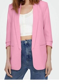 Blazer rosa con una vestibilità su misura, tessuto leggero, apertura frontale, revers a scialle e maniche a tre quarti; indossato sopra un crop top bianco e jeans denim.