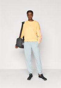 Κίτρινο φούτερ με ροζ ρίγες στα μανίκια, γκρίζες φόρμες και μαύρα αθλητικά παπούτσια. Το μοντέλο κρατάει μια μαύρη τσάντα με λογότυπο.