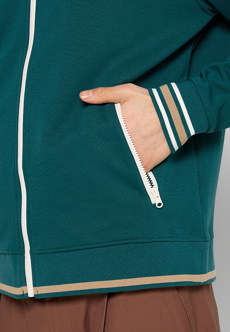 Mano nella tasca laterale di una giacca zip-up turchese con cerniera bianca e polsini a righe, indossata con pantaloni marroni.
