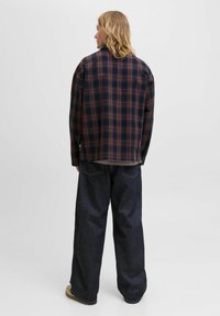 Karierter Overshirt in Marineblau, Braun und Weiß mit einer lockeren Passform, getragen über einem grauen Oberteil. Kombiniert mit lockeren, dunklen Jeans und beigen Schuhen.
