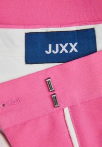 Roza tkanina glatke teksture, s plavom oznakom "JJXX", srebrnim detaljima i bijelim zatvaračem.