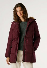 Chaqueta acolchada burdeos con capucha, que presenta un borde de piel sintética, textura acolchada, bolsillos laterales y un cierre frontal con cremallera. Llevada sobre un suéter gris.