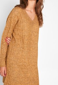 Senapsgul stickad tröja med V-ringning, ribbad textur och långa ärmar, med en avslappnad passform som sträcker sig strax förbi höfterna.