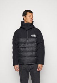 Giacca a vento nera con colletto alto, zip a metà davanti e grande tasca frontale. Presenta una texture trapuntata e un logo sul petto.