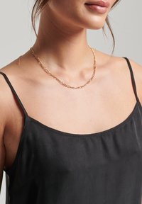 Collana d'oro con design intrecciato e testurizzato, indossata su un top nero con spalline sottili. I materiali sembrano metallici e lucidi.