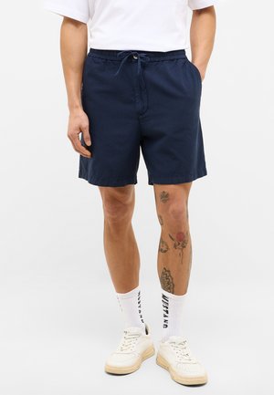 Pantalones cortos de algodón azul marino con cintura elástica, bolsillos laterales y un ajuste relajado, combinados con calcetines blancos y zapatillas.