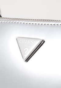 Zilveren, glanzende handtas met een driehoekig zilveren metalen logo met "GUESS U.S.A." en een vraagteken. Met rits afsluiting en bijpassende stiksels.