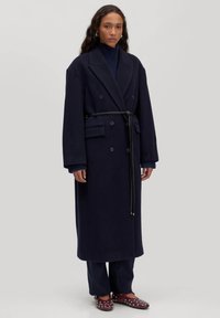 Marineblauwe wollen dubbelbreasted jas met een ceintuur in de taille, reverskraag, zijzakken en verlaagde schouders, gecombineerd met donkere broek en gestifte schoenen.
