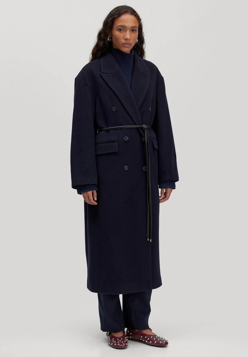 Marineblauwe wollen dubbelbreasted jas met een ceintuur in de taille, reverskraag, zijzakken en verlaagde schouders, gecombineerd met donkere broek en gestifte schoenen.