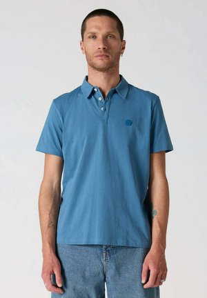 Hombre con pelo corto que lleva una camisa polo azul de manga corta y vaqueros azul claro, de pie frente a un fondo liso de color gris claro.