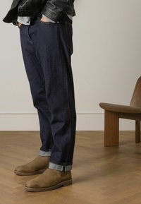 Pantaloni in denim blu scuro con orli risvoltati, abbinati a stivali alla caviglia in suede marrone. Mano dell'uomo in tasca, che indossa una giacca di pelle nera.