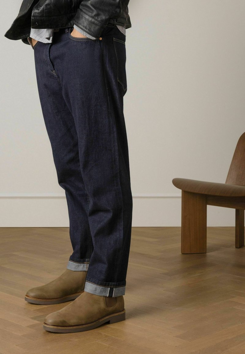 Donkerblauwe denim broek met omgeslagen zoom, gecombineerd met bruine suède enklaarzen. Man's hand in zijn zak, gekleed in een zwart leren jack.