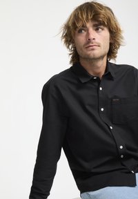 Un jeune homme aux cheveux châtains clairs en désordre regarde ailleurs, portant une chemise noire à boutons avec une poche poitrine et un jean bleu clair.