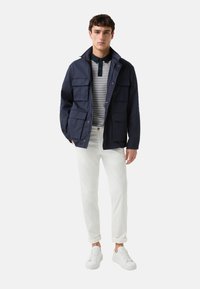 Navyblauwe jas met meerdere voorzakken, een witte en navy gestreepte shirt eronder, witte opgerolde broek en witte sneakers.