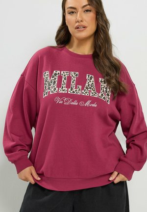 Femme portant un sweat-shirt oversize rose foncé avec "MILAN" en imprimé léopard et texte "Vie Della Moda", debout devant un fond uni.
