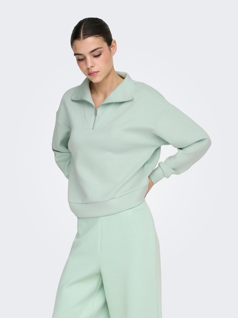 Sweat-shirt vert clair à fermeture éclair avec un col montant et des poignets côtelés, associé à un pantalon large assorti. Tissu doux et texturé.