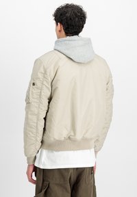 Beige bomber jacket with ribbed cuffs and hem, featuring a gray hoodie underneath. Smooth texture, zip pocket on the sleeve, simple design.