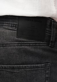 Μαύρο τζιν από τζιν ύφασμα με ένα μαύρο δερμάτινο λογότυπο με την επιγραφή "JACK & JONES" στη μέση, φορεμένο με ένα λευκό πουκάμισο.