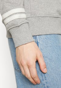 Main reposant à côté de la poche d'un jean bleu clair, portant un sweat à capuche gris avec deux bandes blanches au niveau du poignet.