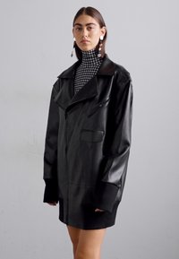 Giacca oversize in ecopelle nera con ampio colletto, tasche con zip e polsini a costine, indossata sopra un dolcevita in bianco e nero a motivo pied de poule.