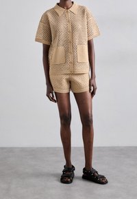 Beige hæklet strikkesæt med en kortærmet knap-op bluse med to lommer og matchende shorts, med en løs pasform og tekstureret design.