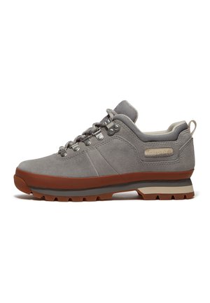 Scarpa da trekking in pelle scamosciata grigia con suola in gomma marrone, occhielli in metallo, collarino imbottito e linguetta in tessuto su sfondo bianco.