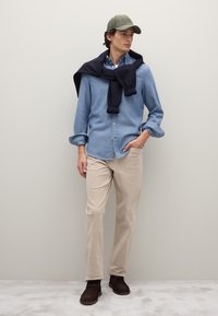 Camicia azzurro chiaro con bottoni, maglione blu navy sulle spalle, pantaloni di velluto a coste beige e stivali marrone scuro. Il look è completato da un berretto verde oliva.