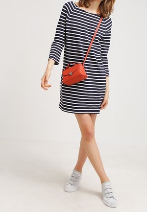 Kobieta w granatowo-białej sukience w paski z rękawami do łokcia, białe sportowe buty i mała czerwona torebka na ramię.