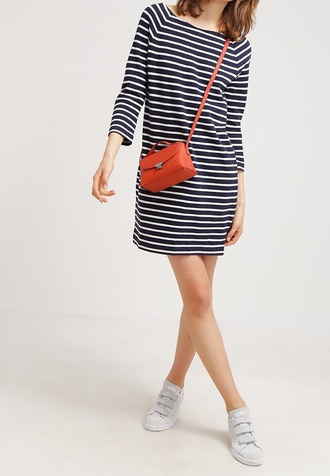 Femme portant une robe rayée bleu marine et blanche à manches trois-quarts, des baskets blanches et un petit sac rouge porté en bandoulière.