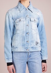 Jasnoniebieska kurtka dżinsowa z klasycznym kołnierzem, przetarciami, dwoma przednimi kieszeniami, srebrnymi guzikami i czarnymi ściągaczami na mankietach.