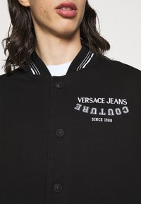 Černá bomber bunda s bílým proužkovaným žebrovaným límcem, s vyšívaným textem "Versace Jeans Couture" a knoflíky vpředu.